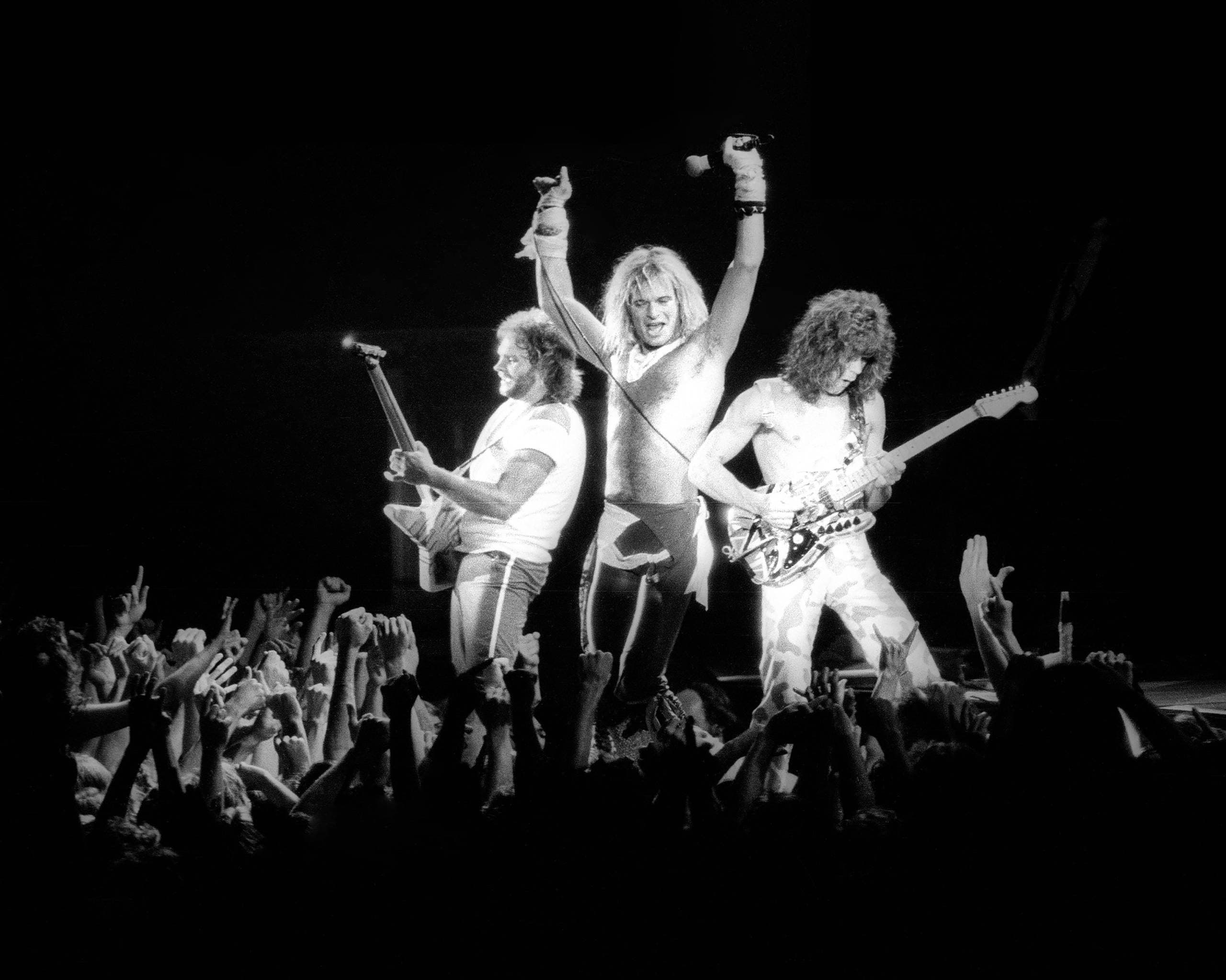 Three musicians on stage, one with a guitar, another raising a mic, fans reaching toward them.