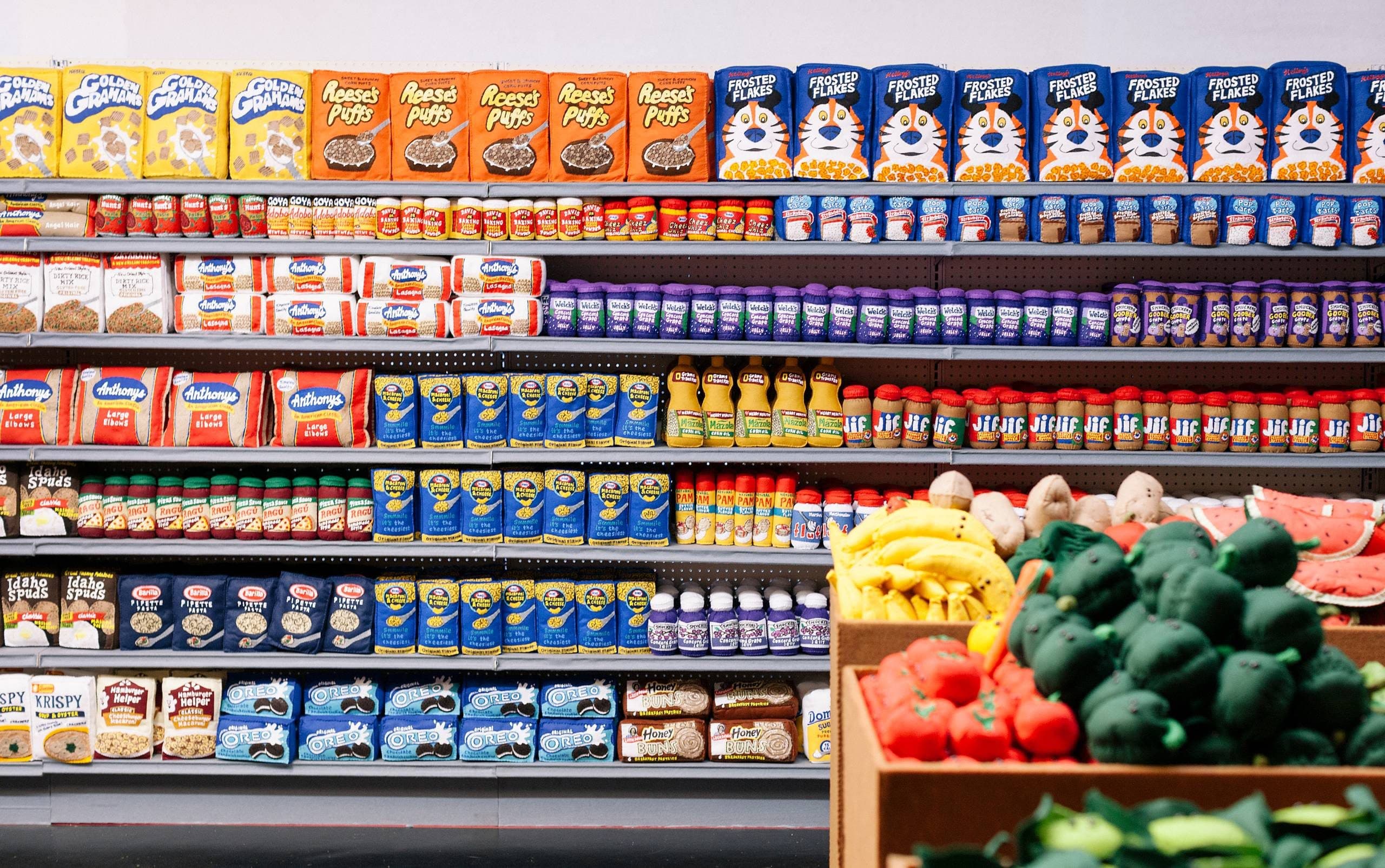 Grocery store shelves stocked with cereals, pasta, canned goods, sauces, and plush toy fruits and vegetables.