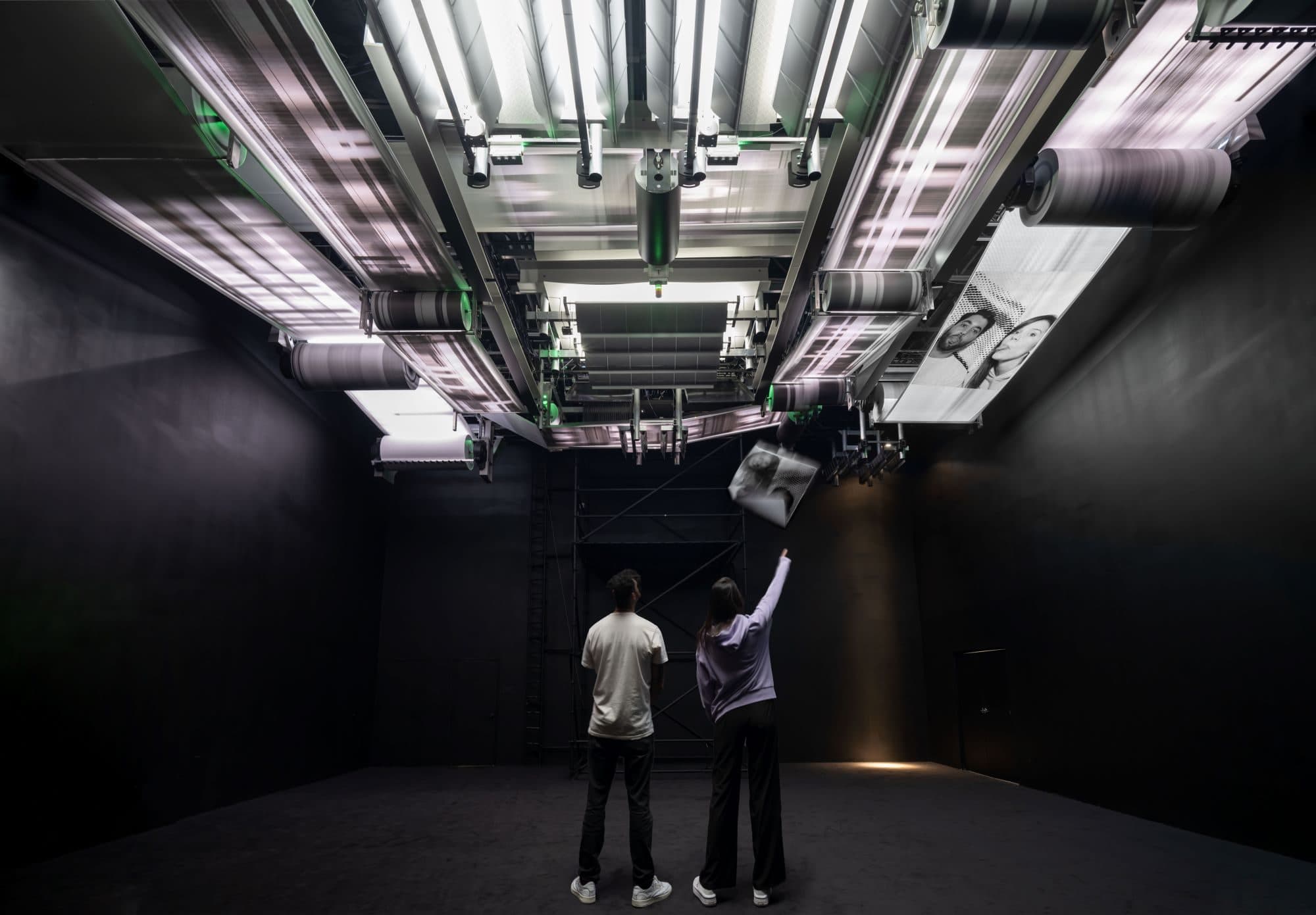 Individuals examining a ceiling-mounted printing system with rollers in a dark room.